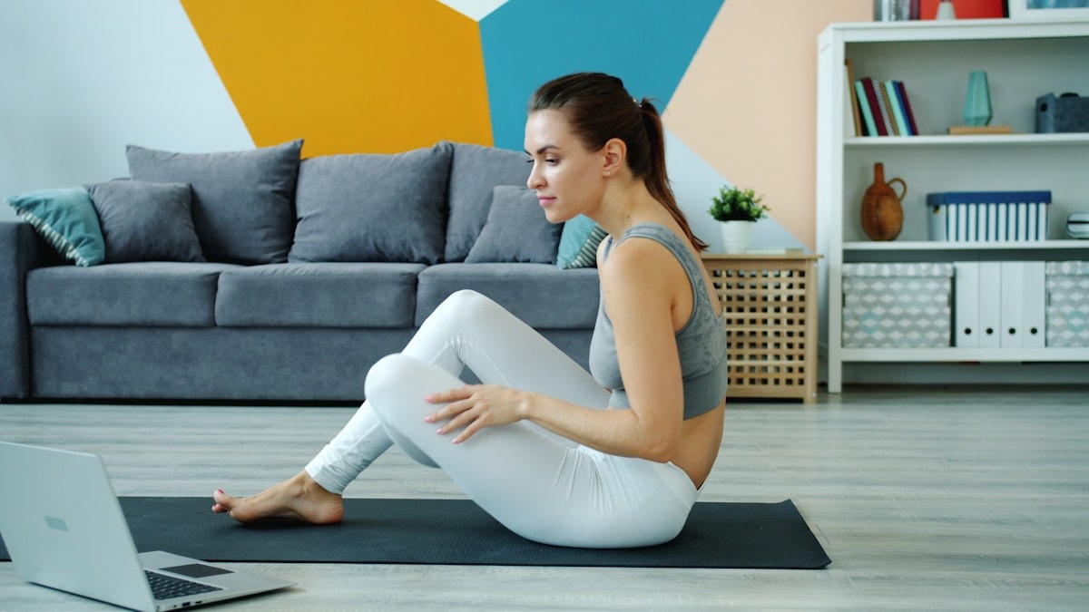 Young lady in top and leggings is using laptop to learn yoga asanas watching tutorial exercising at home on mat. Modern technology and millennials concept.